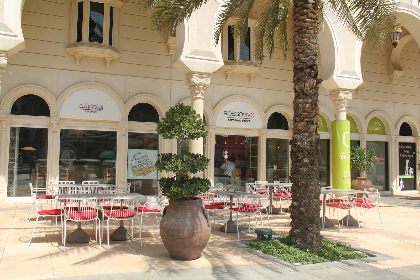 Exterior area fo Rossovivo Sharjah. There is a palm on the foreground, behind the are white chairs with red pillows.